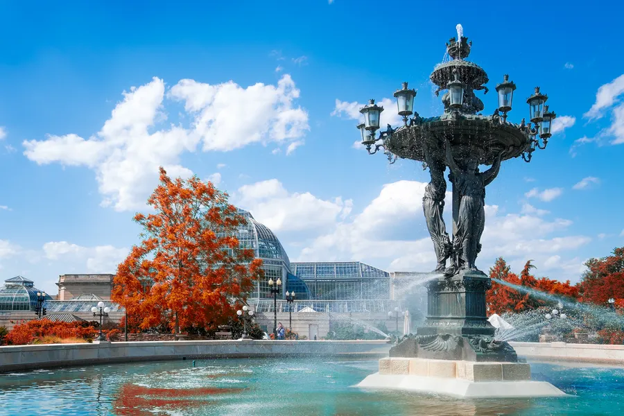 The ornate Bartholdi Fountain in Bartholdi Park in Washington, D.C., photographed in autumn. The dark cast-iron fountain features three classical female figures supporting an upper basin adorned with decorative gas-style lanterns, with water cascading into a large turquoise pool below. Vivid orange and red fall foliage frames the scene on both sides. In the background, the large glass dome of the U.S. Botanic Garden Conservatory is prominently visible against a bright blue sky with puffy white clouds.