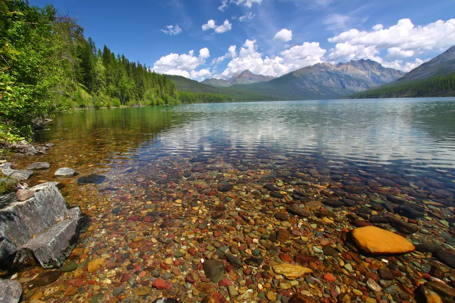 The crystal-clear shallow waters of Kintla Lake reveal a colorful mosaic of multicolored pebbles on the lakebed, with a dense evergreen shoreline and mountain peaks reflected in the calm surface under a partly cloudy sky.