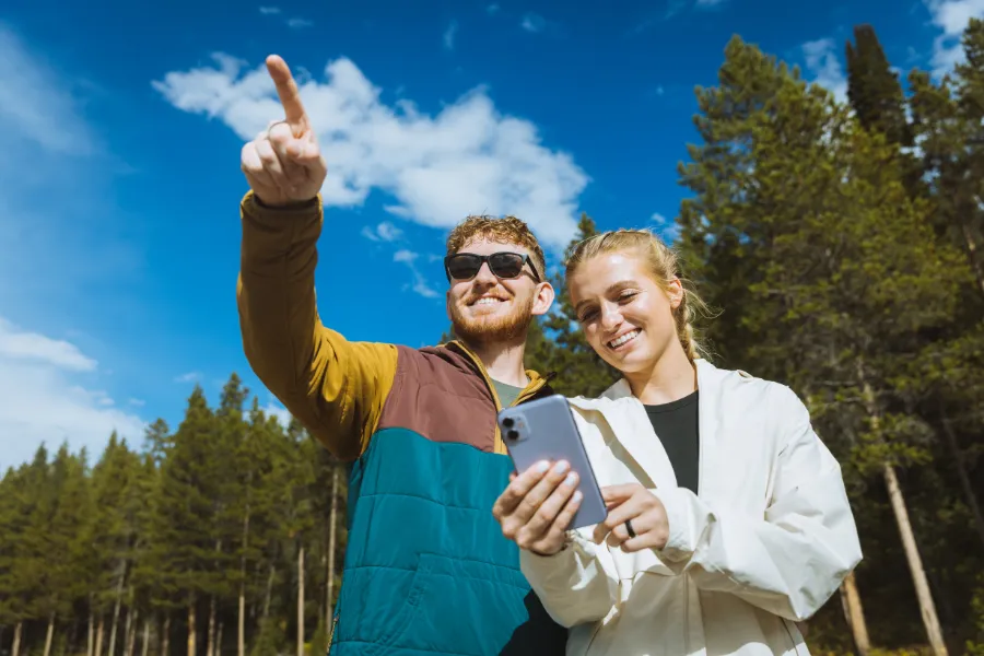 Two travelers stand in a forest clearing smiling at a phone, while one points toward something in the distance under a bright blue sky.