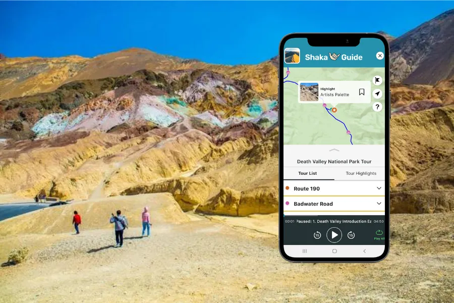 Smartphone displaying the Shaka Guide app over Death Valley’s colorful badlands, with layered mineral-streaked hills and visitors walking below.