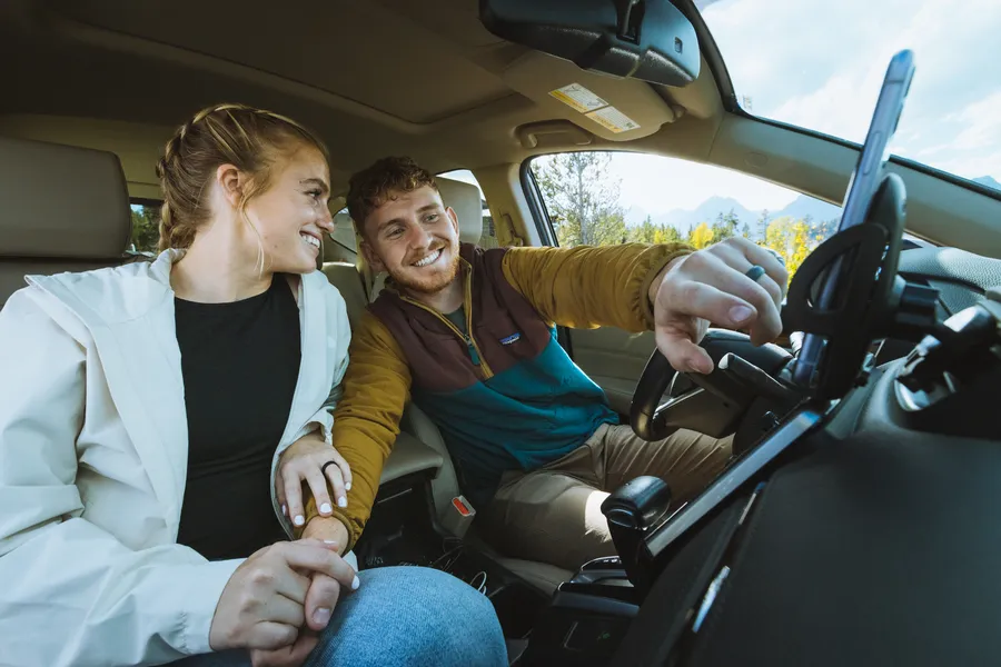 Two travelers smile inside a car while adjusting a mounted smartphone, enjoying the drive together.