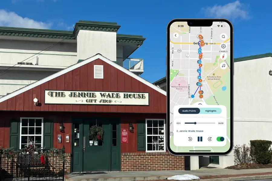 A promotional composite image featuring the exterior of The Jennie Wade House Gift Shop in Gettysburg, Pennsylvania — a red-painted wooden building with dark green doors, white-trimmed windows, a Christmas wreath, and a sign reading "The Jennie Wade House — Gift Shop." Overlaid is a smartphone displaying the Shaka Guide self-guided audio tour app, showing a map of downtown Gettysburg with a vertical blue route and numbered tour stops. The app screen shows stop 2, titled "Jennie Wade House," with audio playback controls and "Audio Points" and "Highlights" tab options.