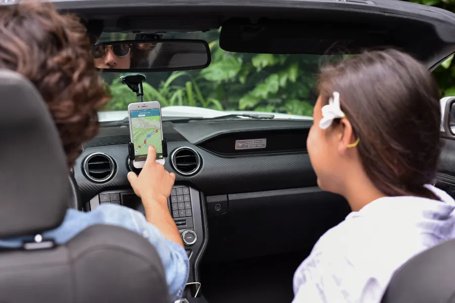 Two travelers sit in a convertible in a lush tropical setting, using a smartphone navigation app mounted on the dashboard.