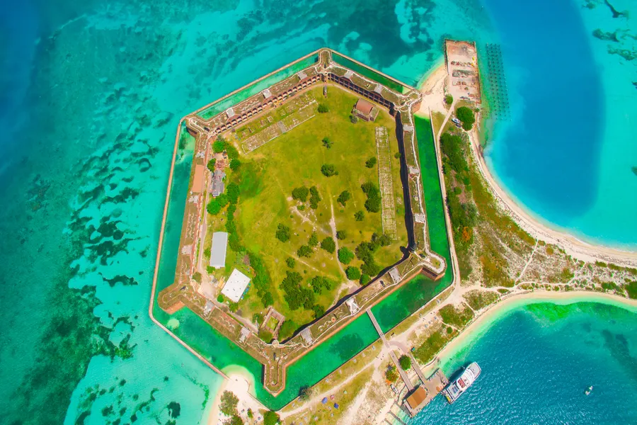 An aerial view of the massive hexagonal Fort Jefferson rises from the vivid turquoise waters of Dry Tortugas National Park, surrounded by a moat, sandy beaches, and coral reefs with a ferry docked at its entrance.