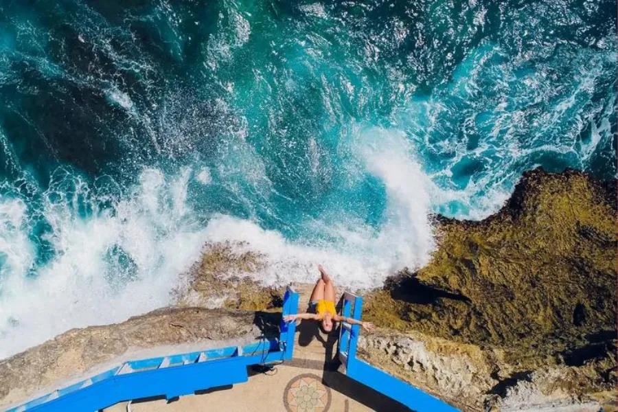 A person relaxes on a bright blue seaside platform above crashing turquoise waves along a rocky cliff edge.