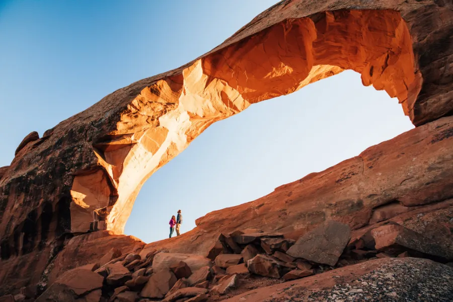 Two hikers stand beneath a massive sandstone arch glowing in warm light above a rocky desert slope.