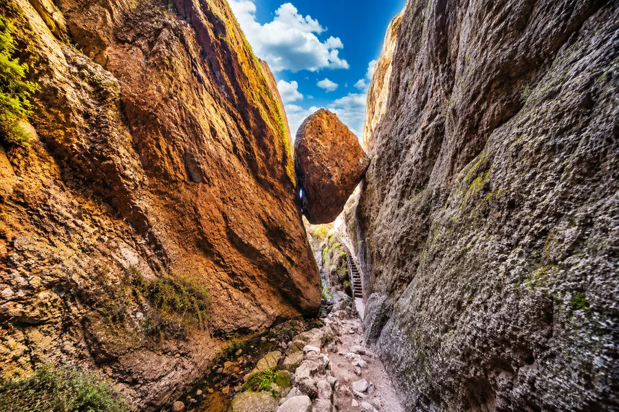 A narrow trail winds through a dramatic rock canyon in Pinnacles National Park, where a massive boulder is wedged high between towering cliff walls. Sunlit orange and gray rock faces rise steeply on either side, while a staircase with a handrail climbs beneath the suspended rock formation. The rugged passage is scattered with stones and small patches of greenery, creating a striking natural scene beneath a bright blue sky dotted with white clouds.