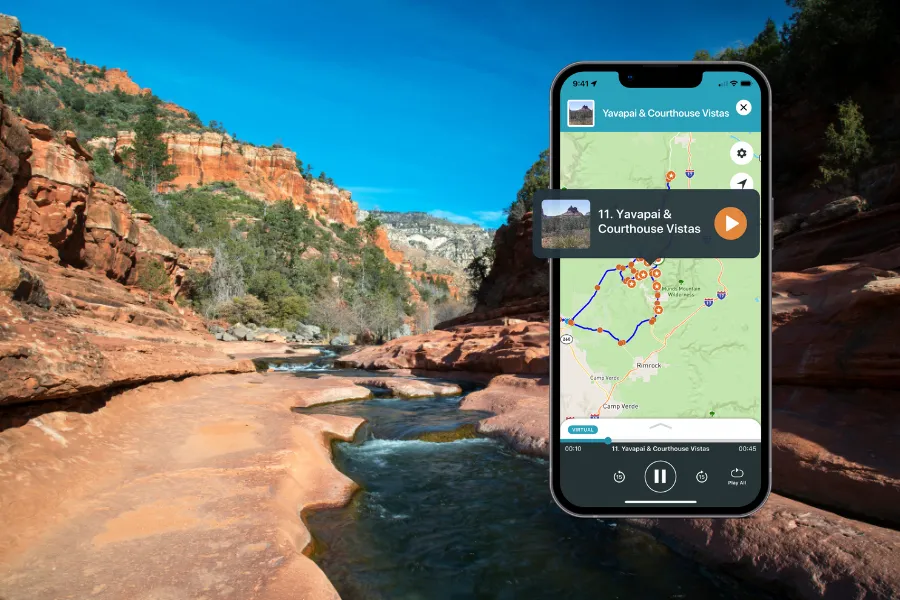 Smartphone displaying a tour app over a red rock canyon stream with flowing water, sandstone cliffs, and a clear blue sky.