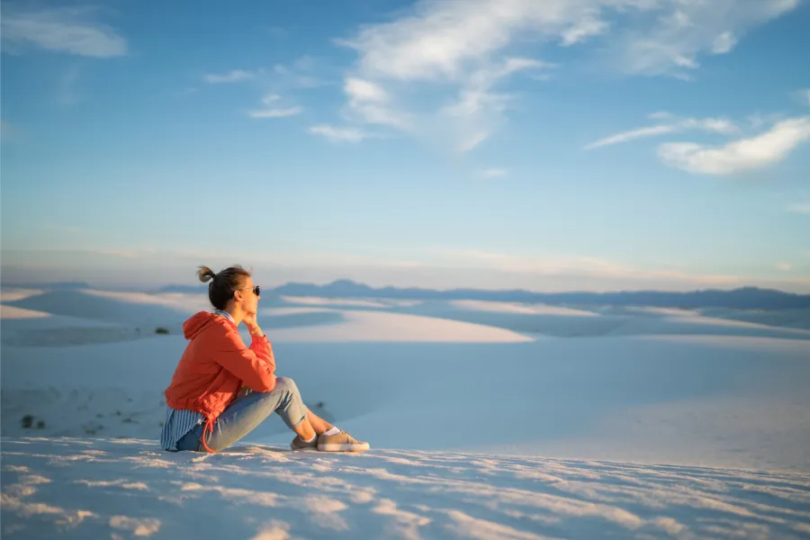 Traveler sits on a white sand dune overlooking rolling desert ridges beneath a soft blue sky.