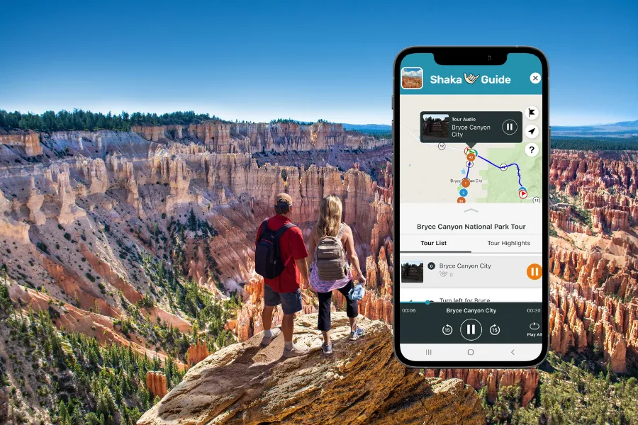 Smartphone displaying the Shaka Guide app over a Bryce Canyon overlook with two hikers standing above a vast landscape of red rock hoodoos.
