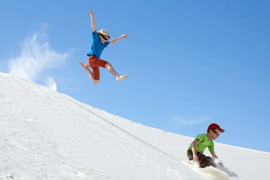 Two children play on bright white sand dunes under a clear blue sky, with one jumping while the other slides down the slope.