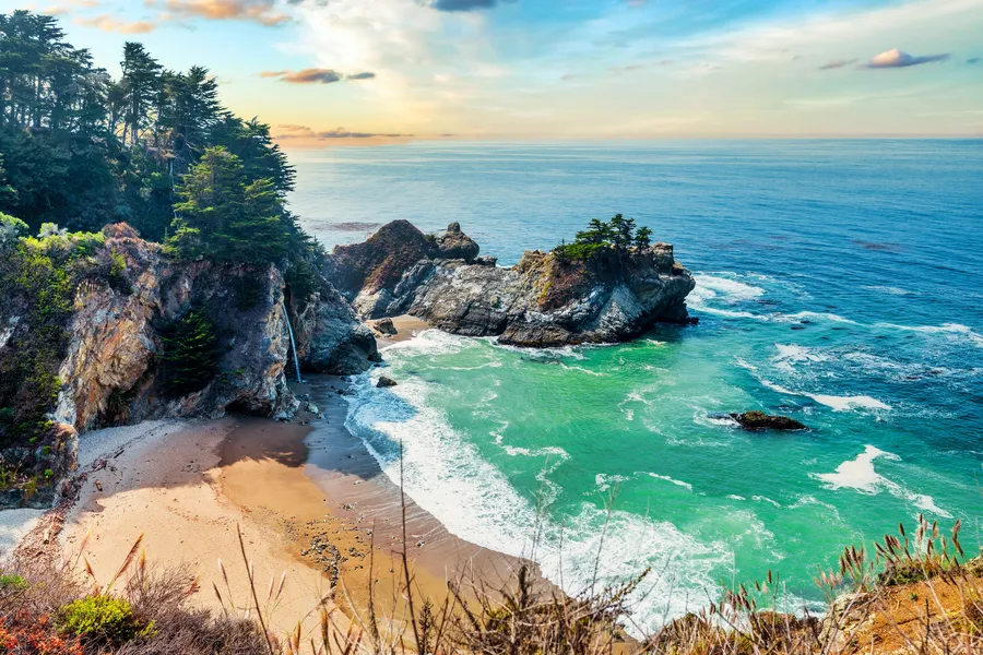 McWay Falls cascades from a coastal cliff onto a secluded sandy cove along California’s Big Sur coastline. Turquoise-green ocean water curls into the beach, while rugged rock formations topped with windswept cypress trees rise from the shoreline. The scene is bathed in soft golden light, highlighting the contrast between the emerald sea, pale sand, and dramatic cliffs.