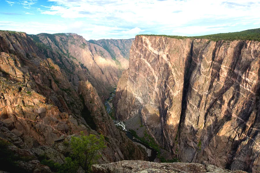 Sheer, near-vertical walls of dark gneiss and schist streaked with pale pegmatite veins plunge dramatically into the narrow gorge of Black Canyon of the Gunnison National Park, with the Gunnison River visible as a thin ribbon far below.