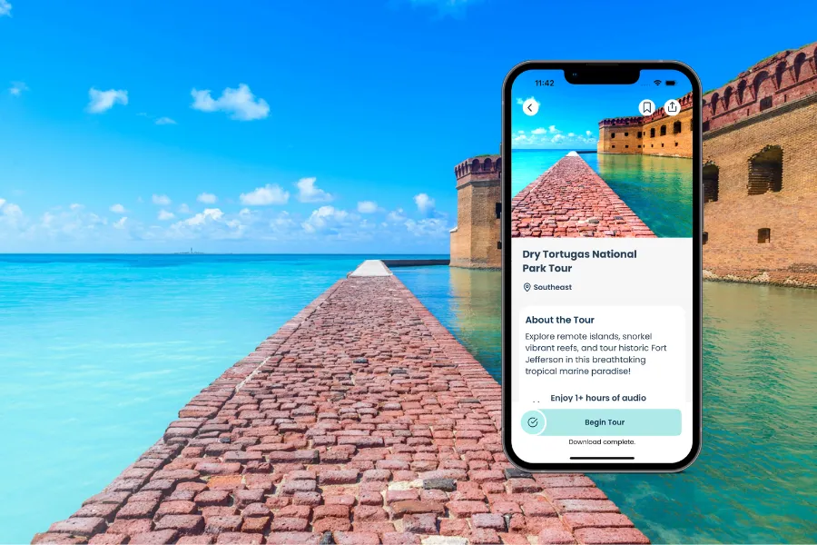 Smartphone screen displays Dry Tortugas National Park Tour app with moat wall in the background, surrounded by turquoise waters and historic brick fort.