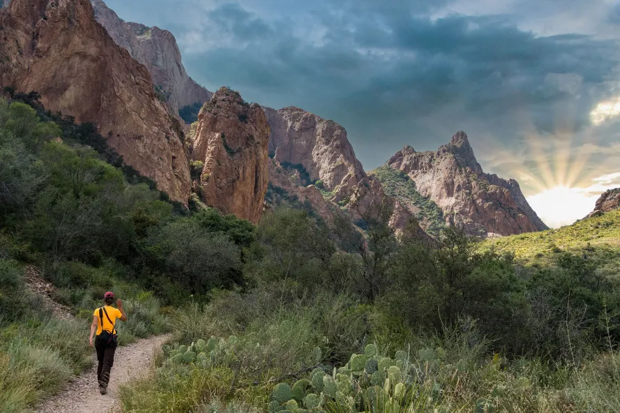 Hiker walks along a desert trail through a rugged mountain canyon, with dramatic clouds and sun rays breaking over the peaks.