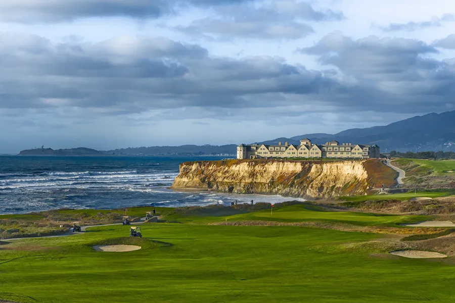 A dramatic coastal landscape overlooks Half Moon Bay, where a large cliffside resort sits atop golden bluffs above the Pacific Ocean. In the foreground, rolling green fairways of a seaside golf course stretch toward the shoreline, dotted with golf carts and sand bunkers. Dark clouds hang overhead while waves crash along the rugged coast, creating a striking contrast between the manicured greens and the wild ocean scenery.