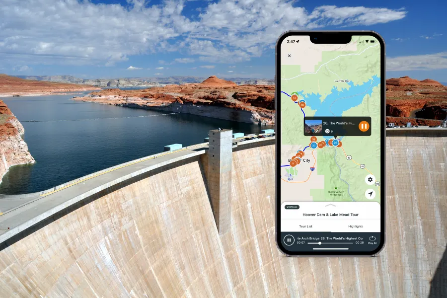 Smartphone displaying a tour app over Hoover Dam, with the curved concrete wall, blue reservoir, and red desert cliffs in the background.