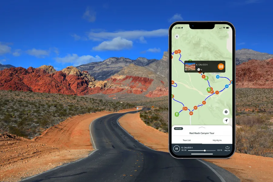 Smartphone displaying a tour app over a winding desert road leading toward layered red rock mountains beneath a vivid blue sky.