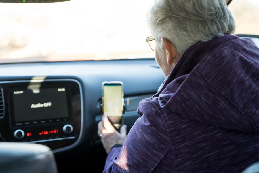 Passenger using a smartphone navigation app mounted inside a car, with the dashboard screen and front windshield visible ahead.