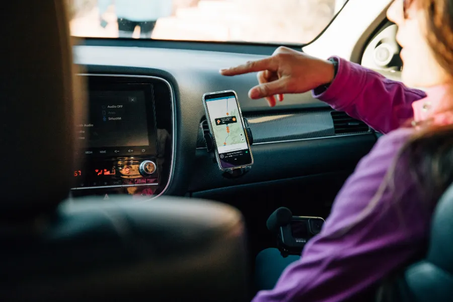 Traveler points toward a smartphone tour map mounted on the dashboard inside a car during a road trip.