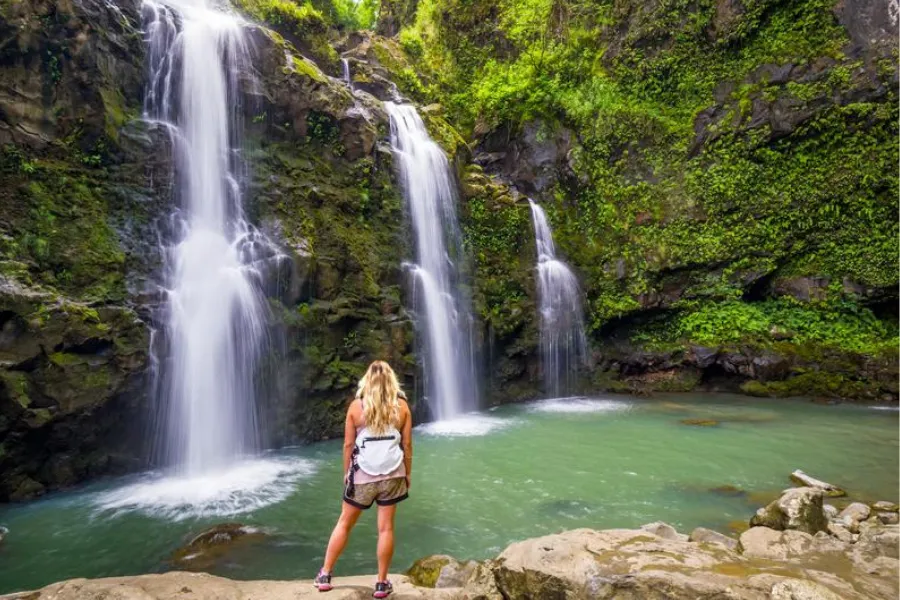 Hiker stands beside a turquoise pool facing a lush multi-tiered waterfall cascading down a moss-covered rocky cliff.