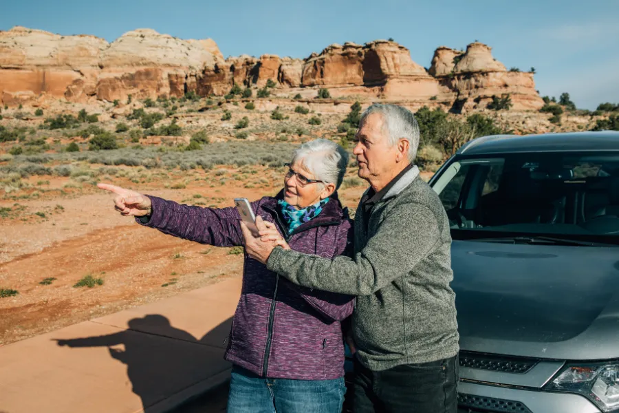 Two travelers stand beside a parked SUV in the desert, with one pointing toward the red rock landscape while holding a phone.