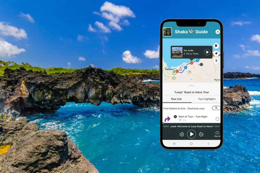 Smartphone displaying the Shaka Guide app over a rocky sea arch with bright blue ocean water under a clear tropical sky.