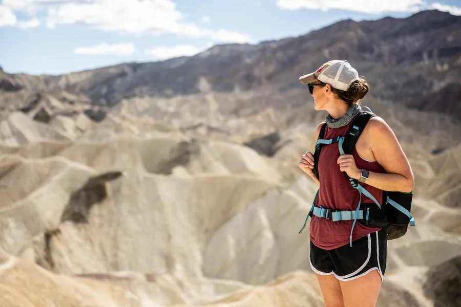 Hiker with a backpack stands at a desert overlook, facing layered badlands and rugged mountains under a bright sky.