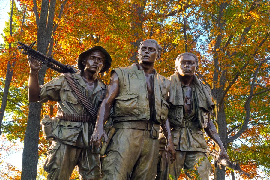 A close-up view of "The Three Soldiers" bronze statue by Frederick Hart near the Vietnam Veterans Memorial in Washington, D.C., photographed against a vivid backdrop of autumn foliage in orange, red, and gold. The highly detailed sculpture depicts three young soldiers in Vietnam-era combat gear standing together — one holding an M16 rifle raised over his shoulder with an ammunition belt across his chest, the center figure gazing upward with an open vest and dog tags, and the third looking to the side with a weapon at his side. The warm fall colors create a striking contrast with the dark bronze figures.