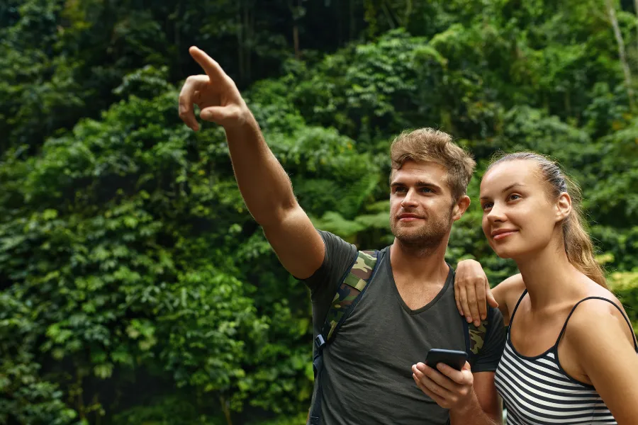Two travelers stand in a lush tropical forest, with one pointing ahead while the other holds a phone and looks on.