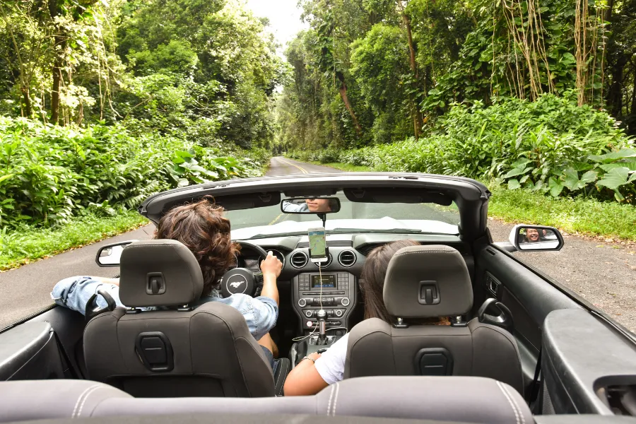 Two travelers drive a convertible along a lush jungle road, with a smartphone navigation app mounted on the dashboard.