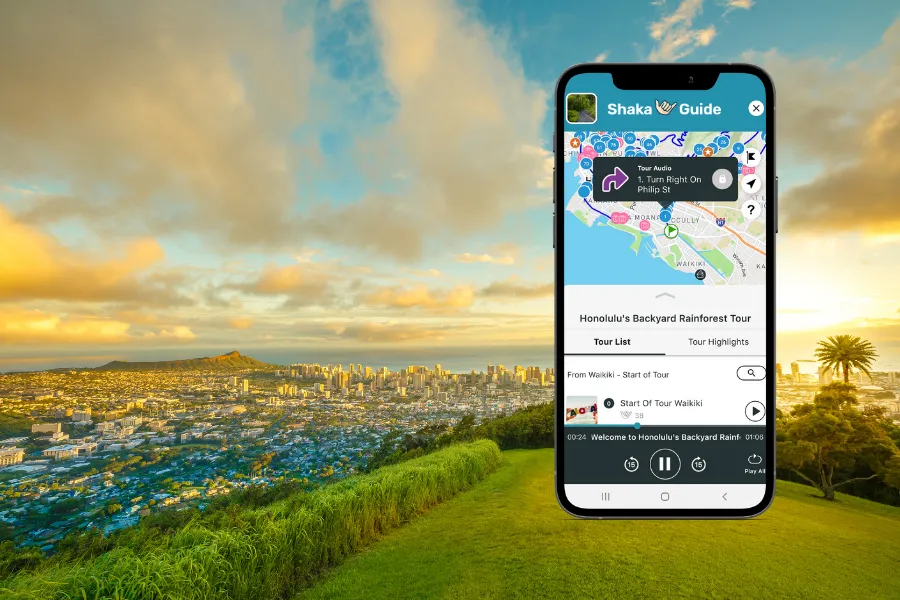 Smartphone displaying the Shaka Guide app over a golden city overlook with green hillside foreground, skyline views, and dramatic sunset clouds above Honolulu.