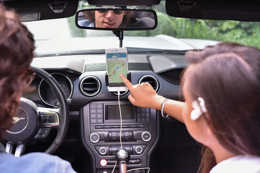 Traveler points to a smartphone navigation app mounted on the dashboard inside a convertible, with the driver visible in the rearview mirror.