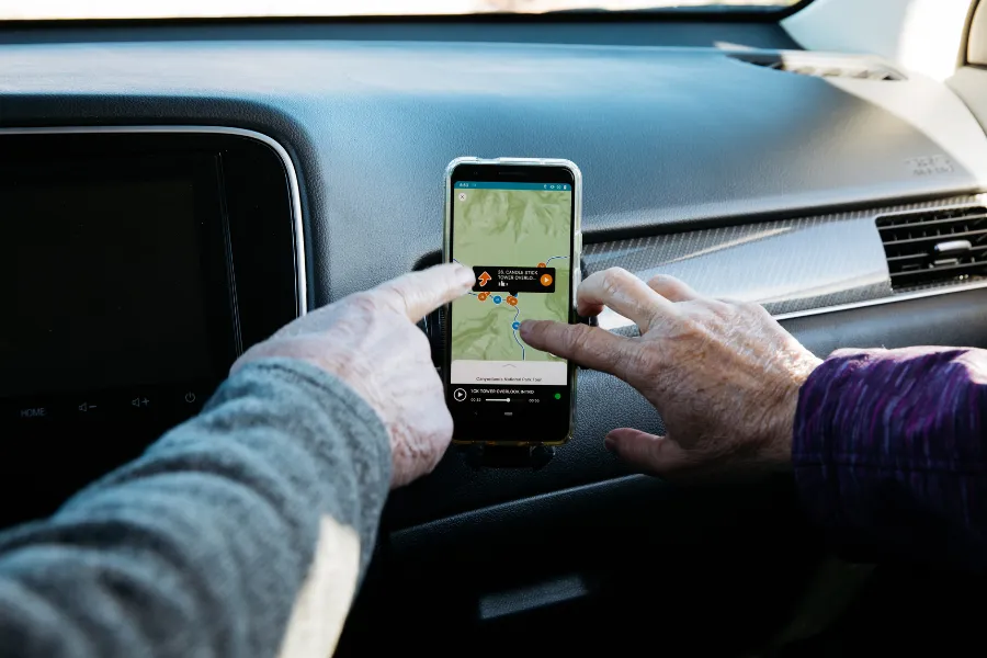 Two hands interact with a smartphone tour map mounted on a car dashboard, with the vehicle interior visible around it.
