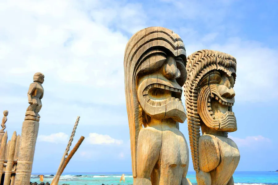 Two carved wooden tiki statues stand by the ocean under a blue sky, with more figures visible in the background.