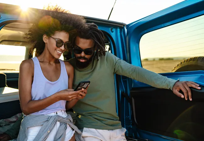 A smiling couple leans on their car and looks at a phone while enjoying a scenic road trip.