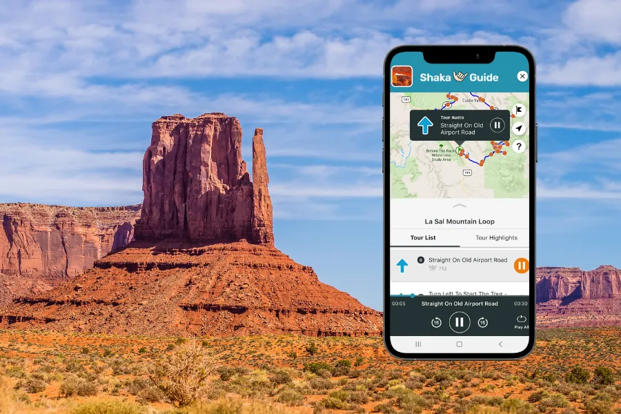 Smartphone displaying the Shaka Guide app over a desert landscape with a towering red rock butte and wide blue sky in the background.