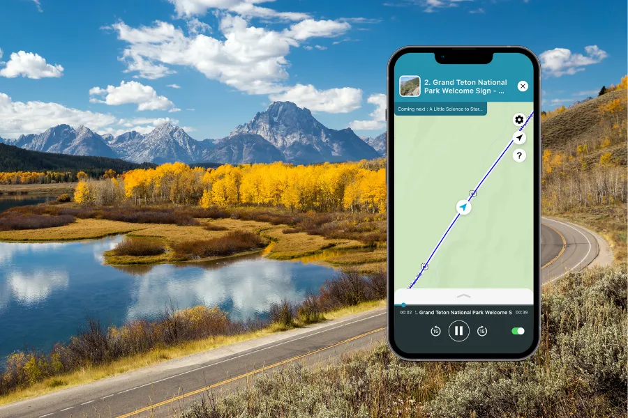Smartphone displaying the Shaka Guide app over a scenic Grand Teton landscape with autumn trees, reflective water, mountain peaks, and a winding road.