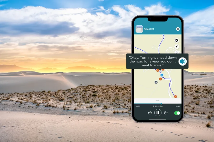 Smartphone displaying the Shaka Guide app over white sand dunes at sunset, with soft desert ridges and distant mountains beneath a glowing sky.