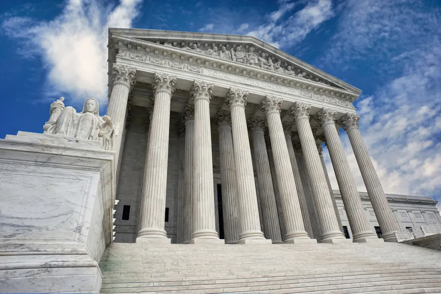 A dramatic low-angle view of the United States Supreme Court building in Washington, D.C., looking up at the grand neoclassical white marble facade. A row of tall fluted Corinthian columns supports an elaborately carved pediment with figurative sculptural relief. The inscription "Equal Justice Under Law" is partially visible along the entablature. Wide marble steps lead up to the entrance, and an allegorical seated marble statue is visible on the left side of the staircase. The building is set against a vivid blue sky with sweeping white clouds.
