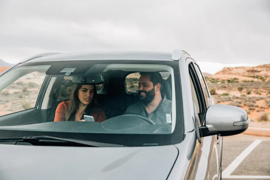 Two travelers sit inside an SUV in a desert landscape, looking at a smartphone while parked under an overcast sky.