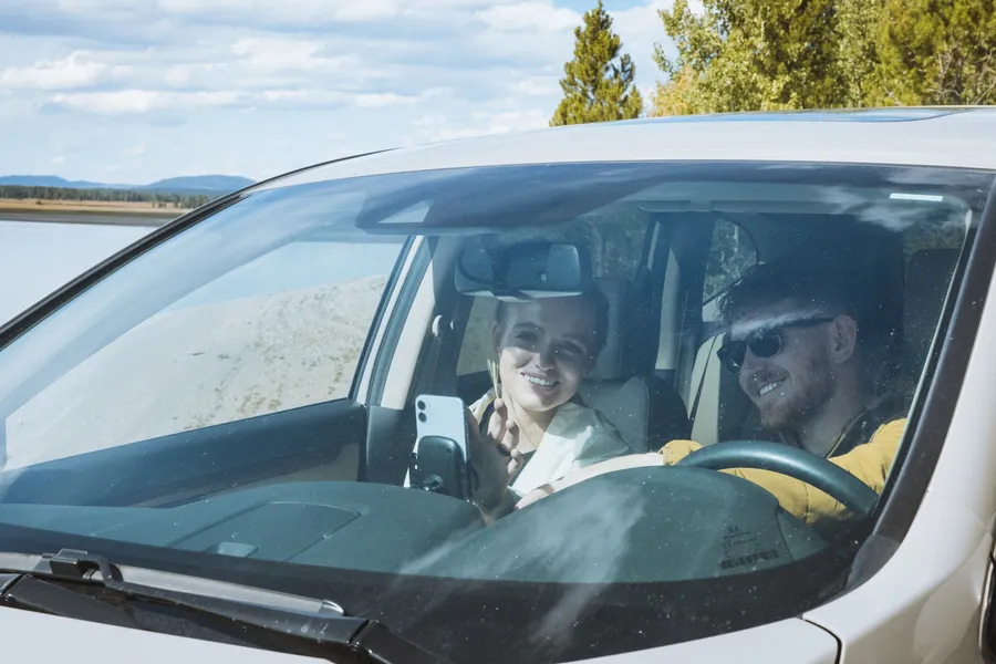 Two travelers smile inside a car by the lakeshore, with a phone mounted on the dashboard and water visible outside.