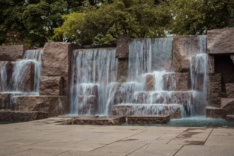 A long-exposure photograph of the multi-tiered waterfall feature at the Franklin Delano Roosevelt Memorial in Washington, D.C. Water cascades smoothly down several levels of large, rough-cut red granite blocks, creating a silky, misty effect. The water collects in a shallow reflecting pool at the base, bordered by a broad granite plaza in the foreground. Dense green trees rise above the granite walls in the background, partially visible against an overcast sky.