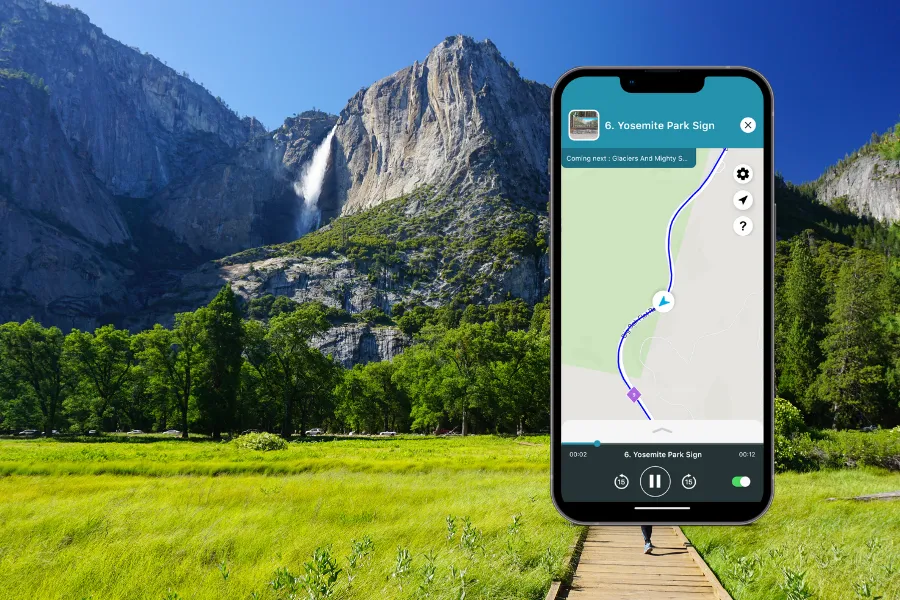 Smartphone displaying the Shaka Guide app in Yosemite Valley, with granite cliffs, a waterfall, green meadow, and boardwalk in the background.