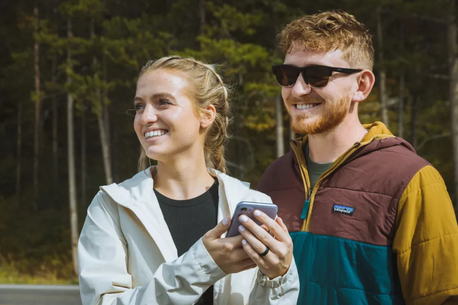 Two travelers smile while holding a phone outdoors, standing in front of a forested backdrop.