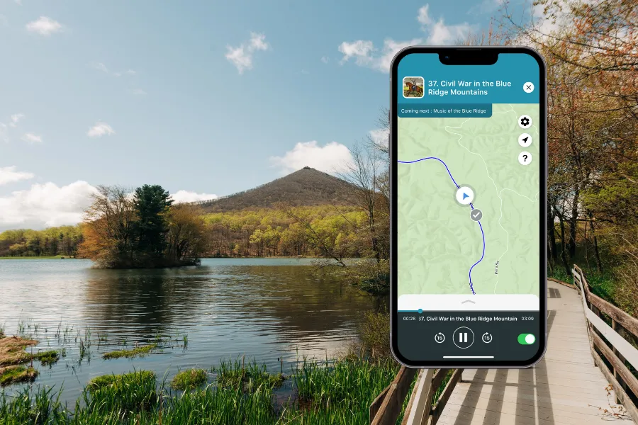 Smartphone displaying the Shaka Guide app over a peaceful lakeside boardwalk with forested hills and mountain views under a clear sky.