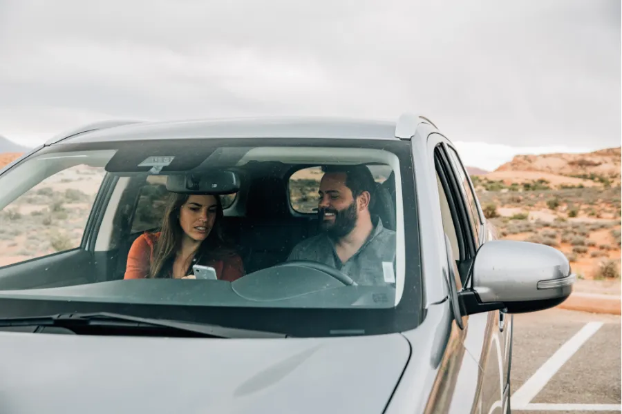 Two travelers sit inside an SUV in a desert landscape, looking at a smartphone while parked under an overcast sky.