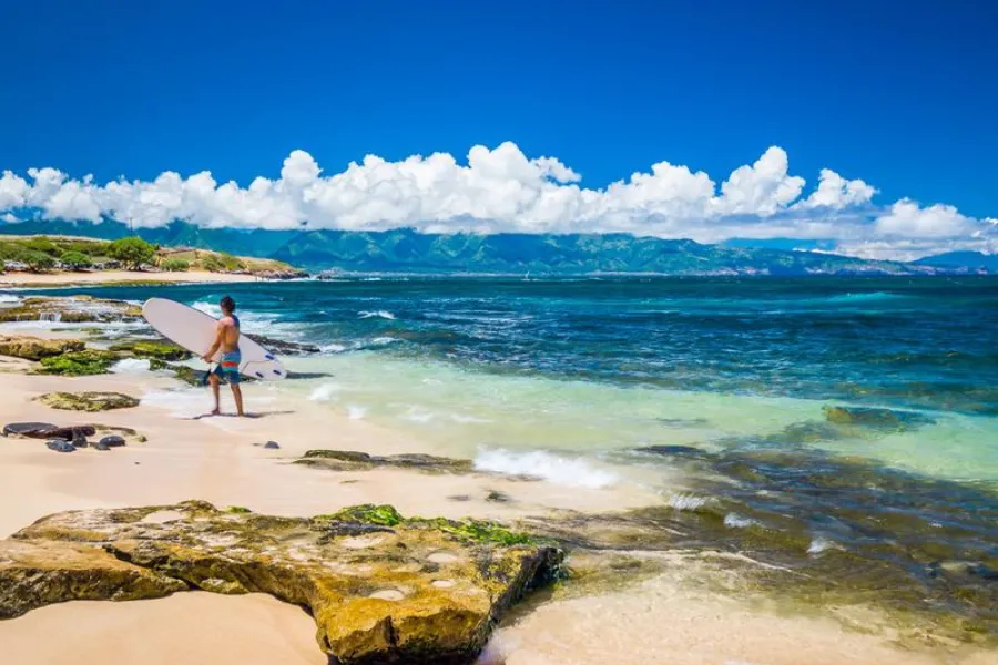 Surfer stands on a sandy beach with a board beside clear blue water, rocky shoreline, and distant mountains under bright clouds.