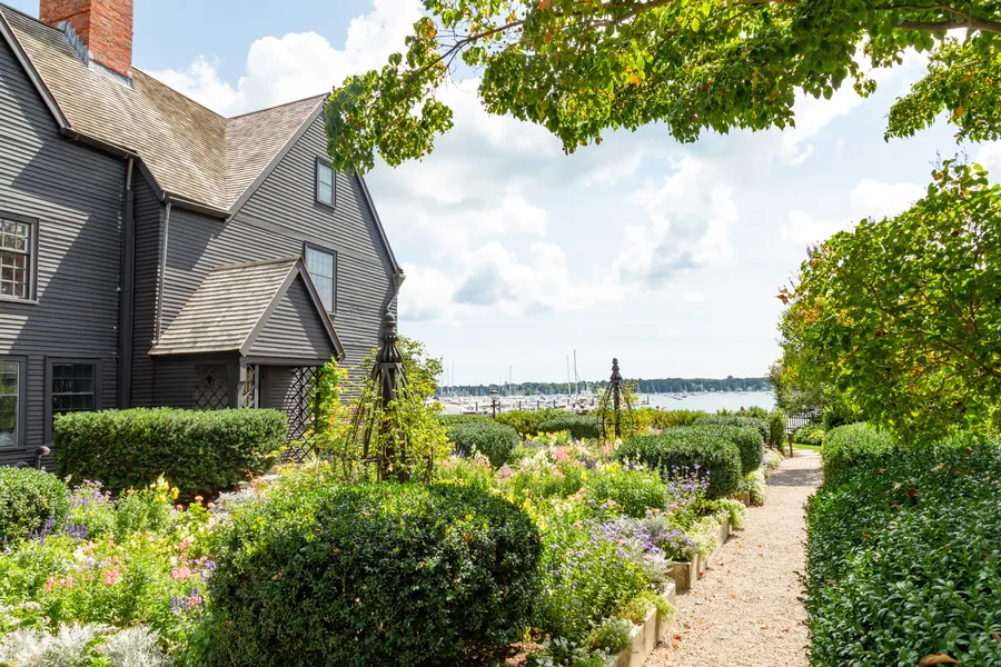 The historic House of the Seven Gables rises beside a lush colonial garden with manicured hedges and colorful flowers, overlooking Salem Harbor with sailboats visible in the background.