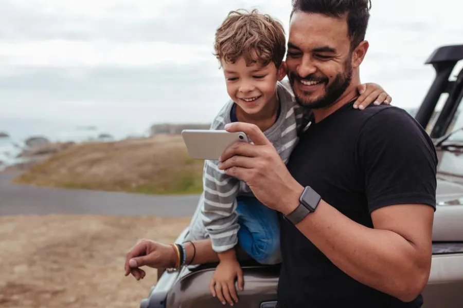 Adult and child smile while looking at a phone beside a parked vehicle near an open coastal landscape.